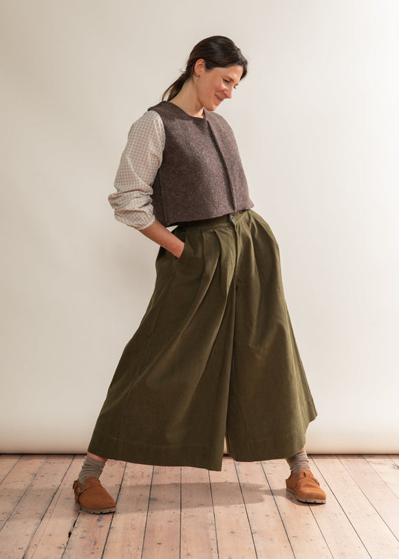 Woman wearing a brown vest and green wide-leg pants standing on a wooden floor.