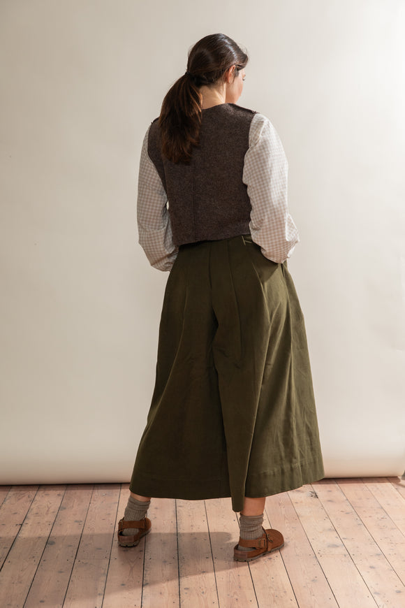 Person wearing a vest, shirt, and wide-leg pants standing against a plain wall.
