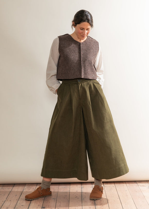 Woman wearing a brown vest, white shirt, and green wide-leg pants against a white wall.