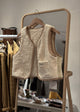 Beige textured vest on a hanger in front of a mirror with clothing rack in the background