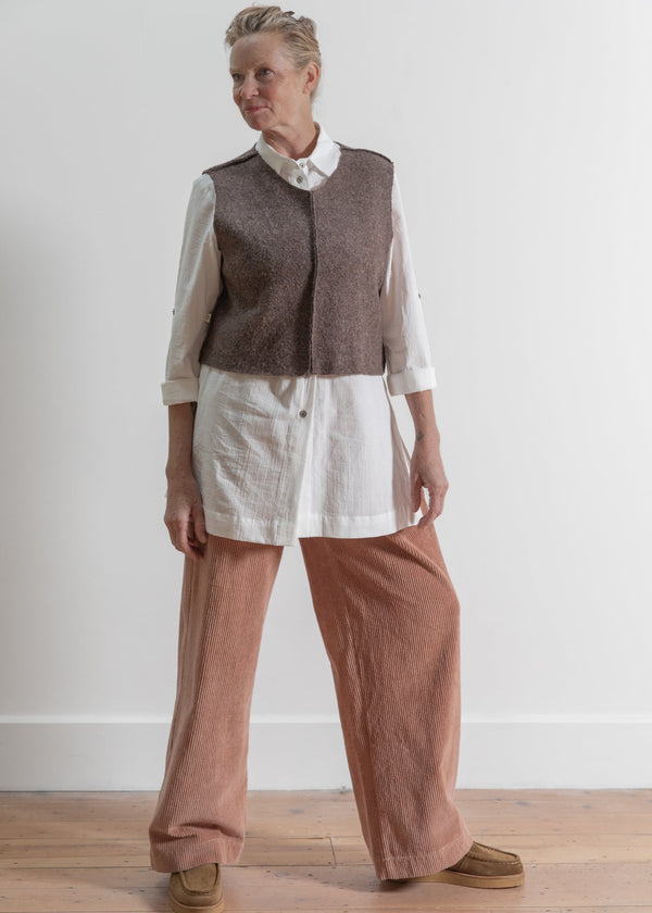 Person wearing a brown vest over a white shirt with light brown pants against a plain background