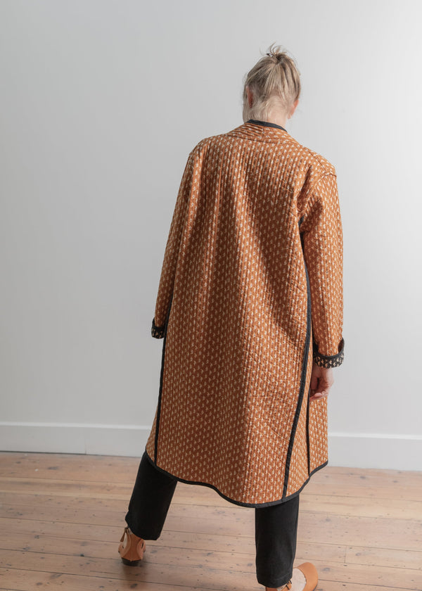 Person wearing a brown patterned coat standing in a room with wooden flooring and white walls.
