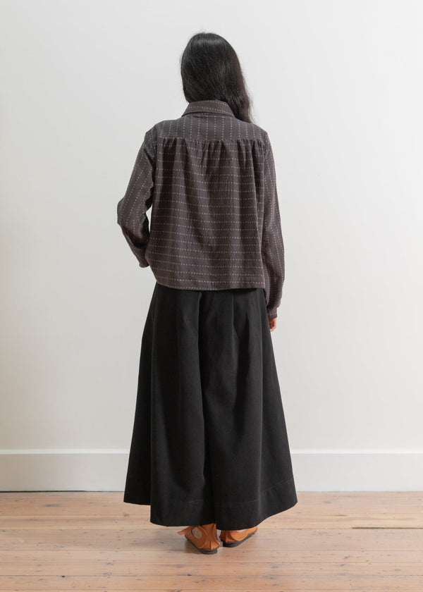 Person wearing a greycheckered shirt and black wide-leg pants standing against a white wall.