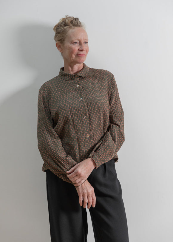 Person wearing a patterned shirt and dark pants against a plain background