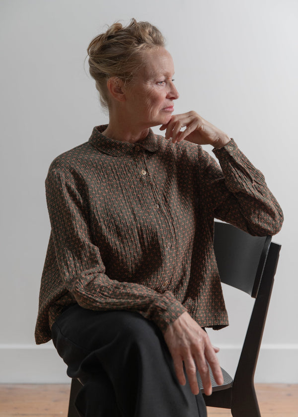 Woman sitting on a chair wearing a brown textured blouse against a plain white wall.