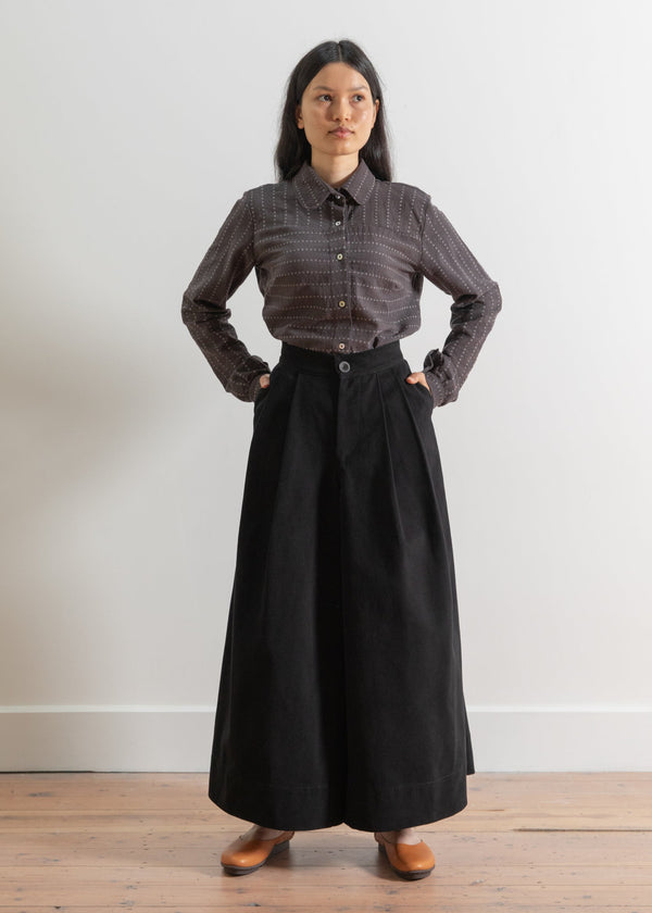 Woman wearing a brown button-up shirt and black skirt standing on a wooden floor with a white wall background.