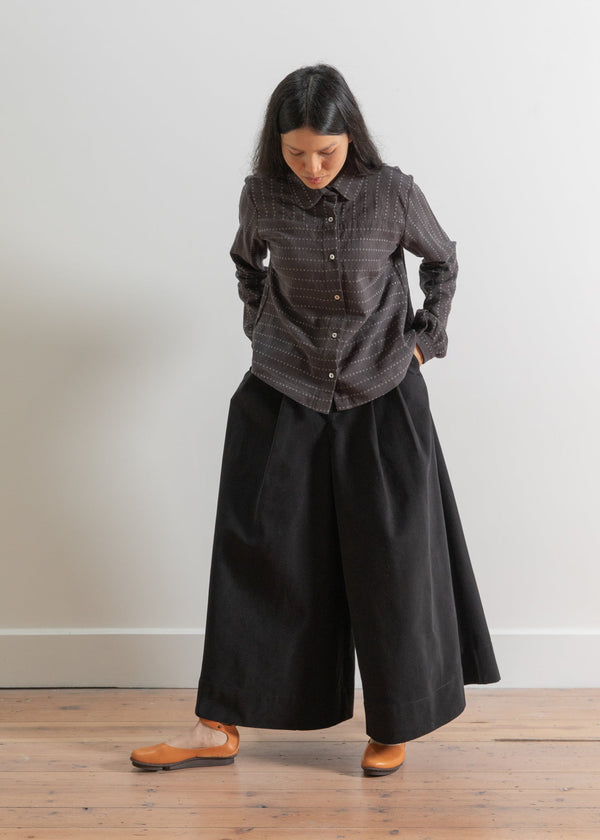 Person wearing a dark plaid shirt and black wide-leg pants against a plain background