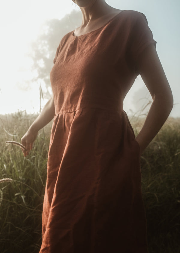 A woman wearing an orange, boxy body, sleeveless, midi dress with drop down sleeves, standing in a field.