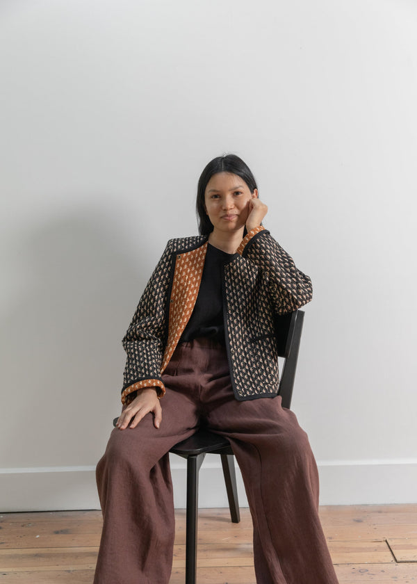 Woman sitting on a chair wearing a patterned jacket and brown pants against a plain background