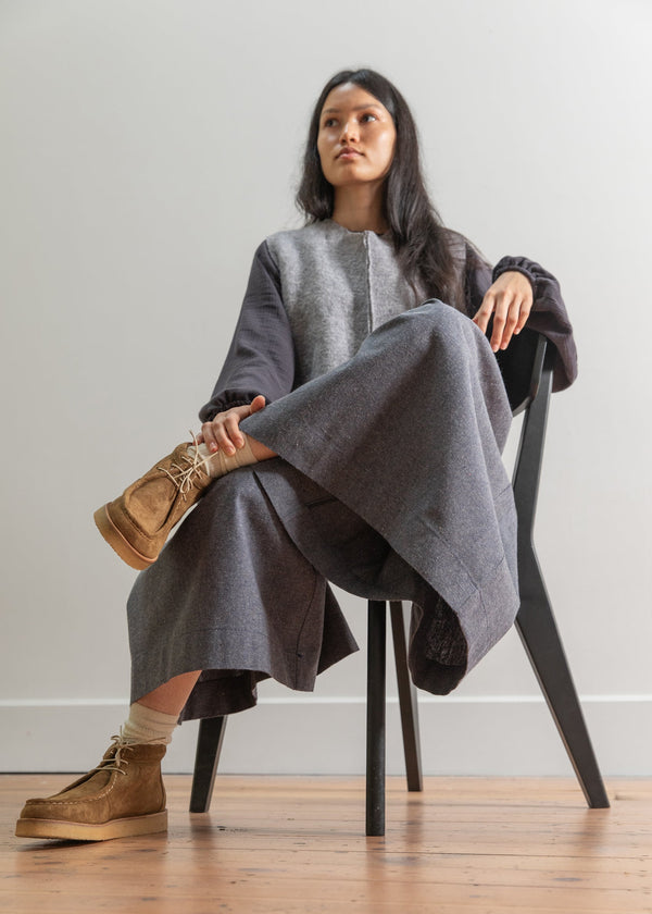 Woman sitting on a chair wearing a gray coat and brown boots in a minimalistic setting.