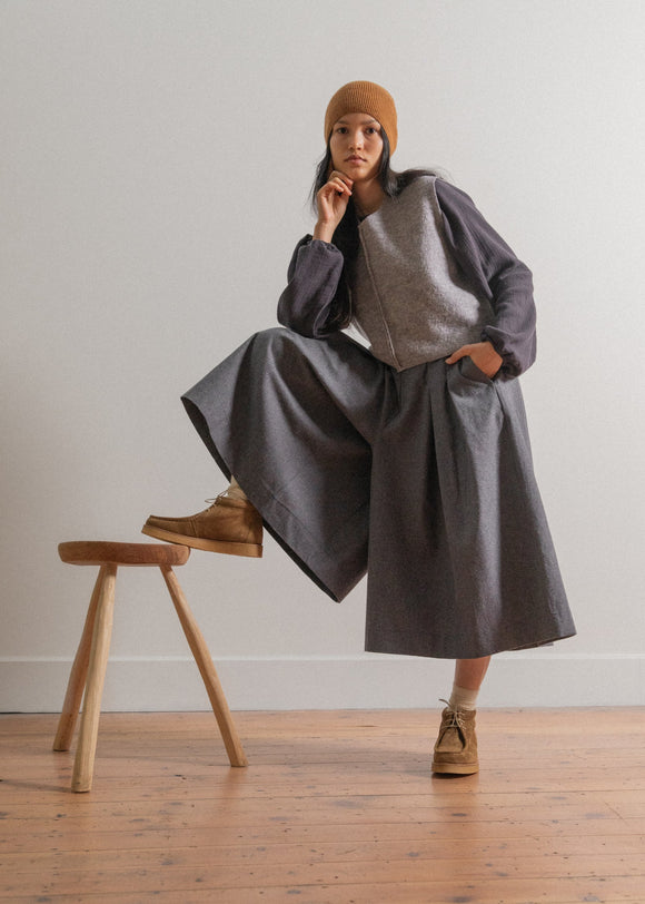 Person wearing a gray outfit with a brown beanie and shoes, sitting on a wooden stool.