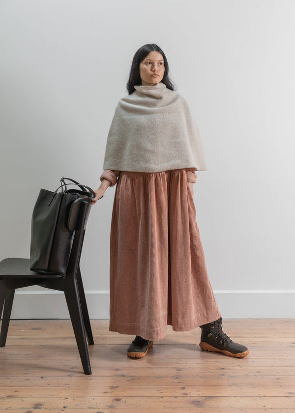 Woman wearing a beige poncho and pink skirt standing in a room with a chair and bag.