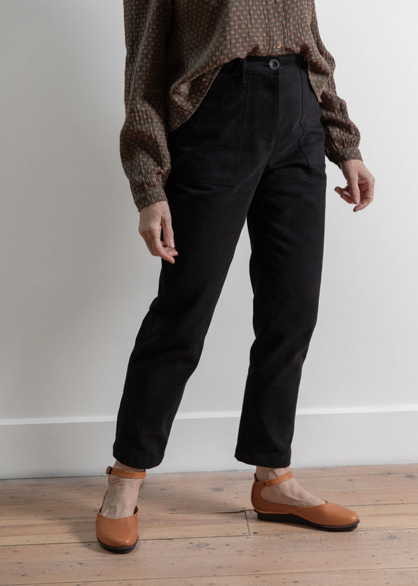 Person wearing black pants and brown shoes on a wooden floor with a white wall background