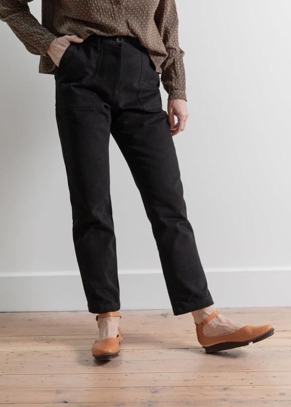 Person wearing black pants and brown shoes on a wooden floor with a white wall background