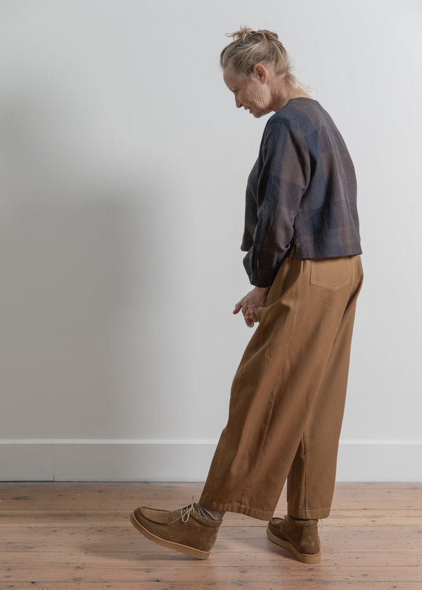 Person wearing a plaid shirt and brown pants standing on a wooden floor against a white wall.
