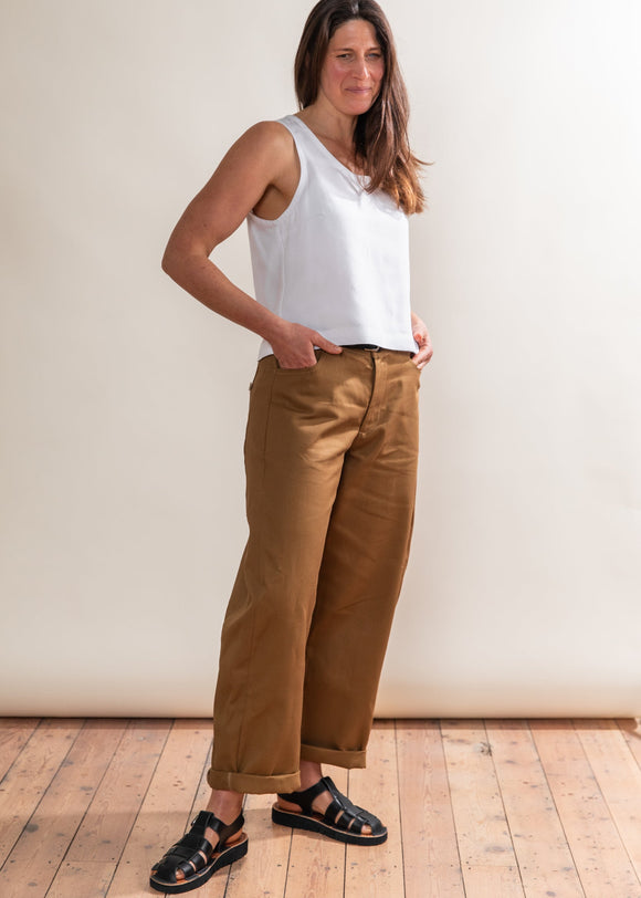 Woman wearing a white top and brown pants standing against a plain wall.