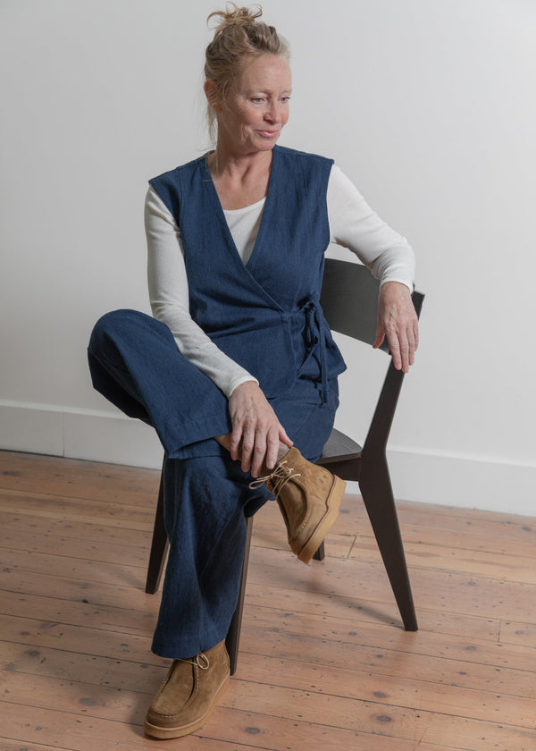 Woman sitting on a chair wearing a blue vest and pants with brown shoes.