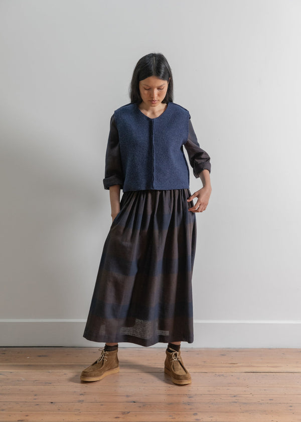 Woman wearing a dark blue top and black skirt standing on a wooden floor against a plain white wall.