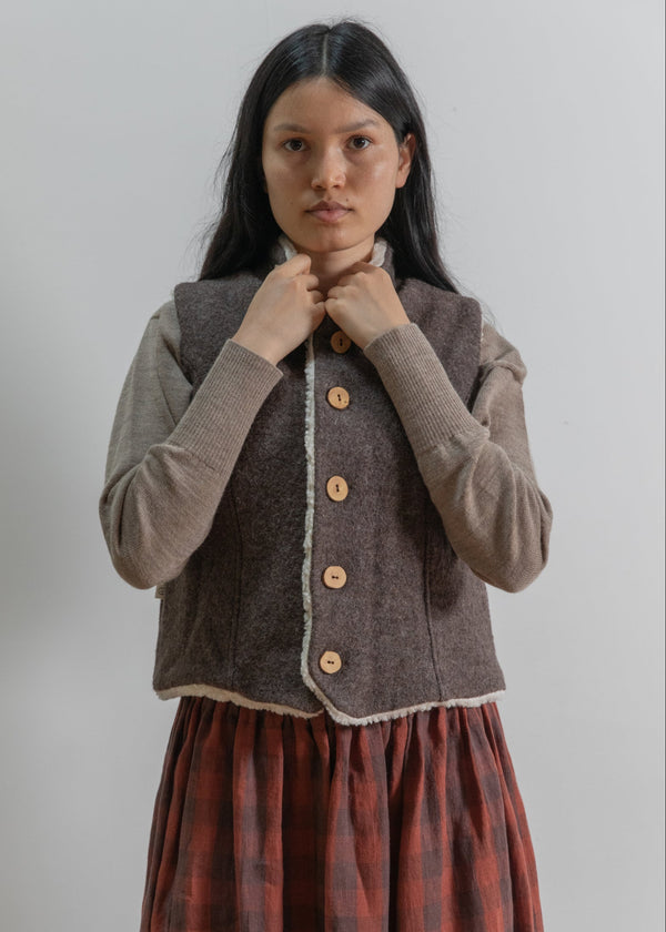 Woman wearing a brown vest over a gray sweater with a red plaid skirt on a plain background