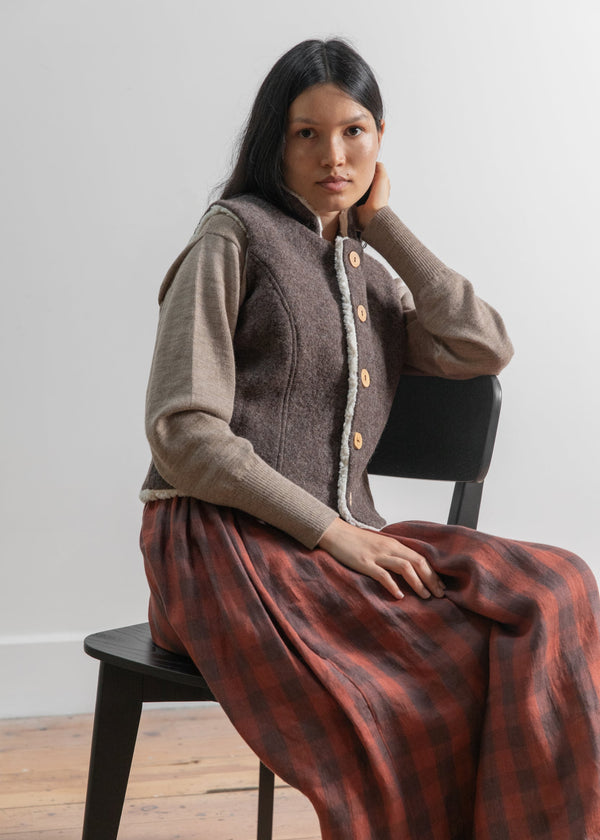 Woman wearing a brown vest and red plaid skirt sitting on a black chair against a white wall.