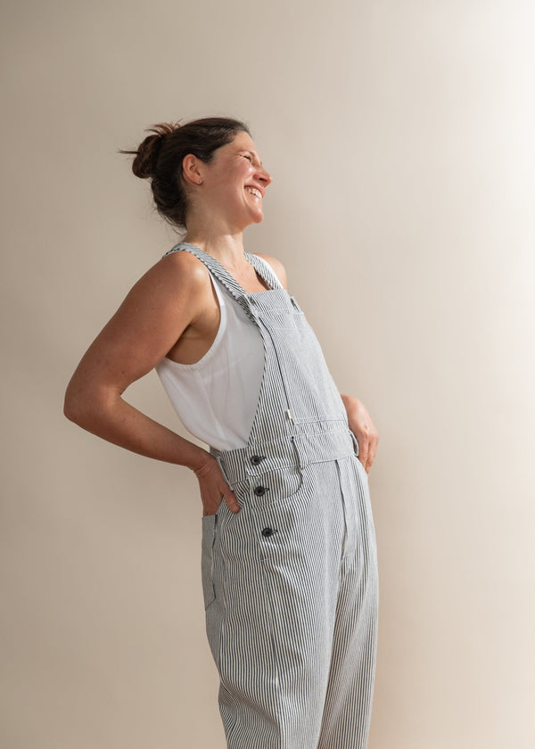 Woman wearing light blue checkered overalls and a white tank top against a plain background