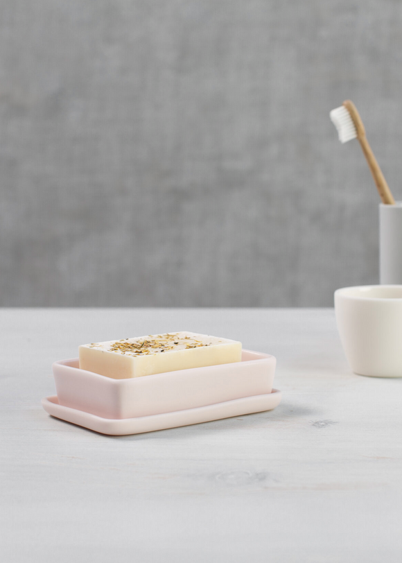A white porcelain soap dish with a pinkish hue, accompanied by a white cup and a small vase, all placed on a grey surface with a textured grey background.