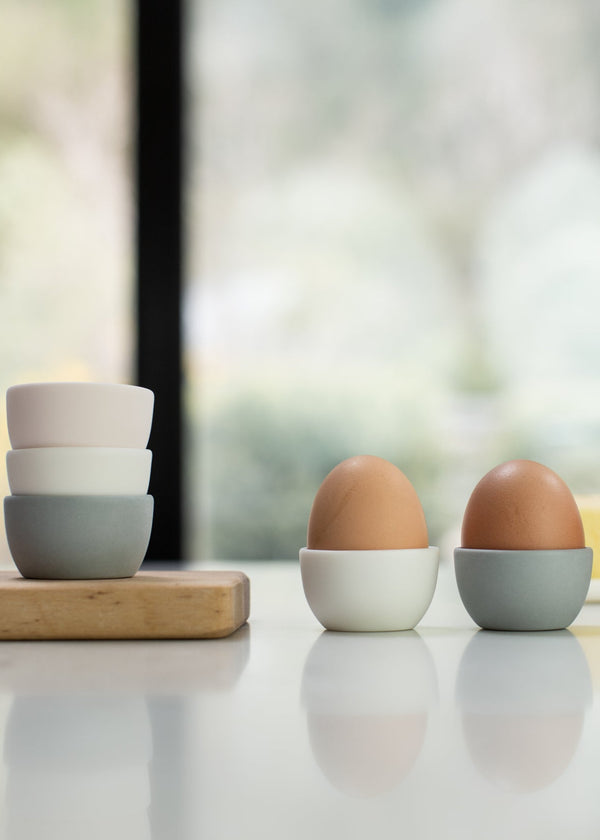Three eggs in small ceramic containers on a light surface with a blurred window background