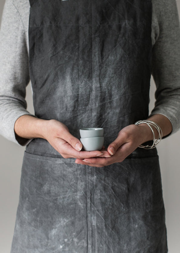 Person wearing a grey apron holding a small white egg cup against a plain background