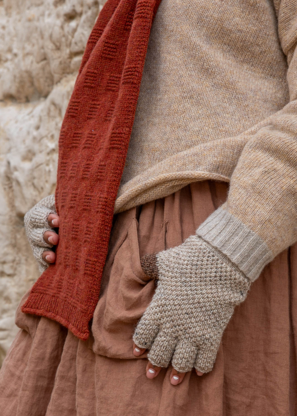 Person wearing a beige sweater, brown skirt, and gray gloves holding a red knitted item against a textured beige background.