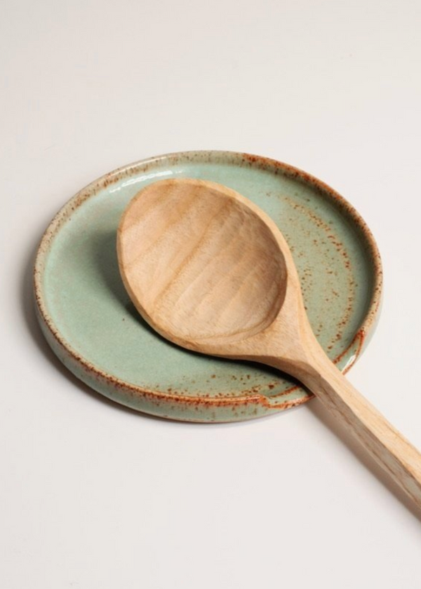 A beige stoneware spoon rest with a satin sea kelp and gloss overlay glaze on a white background, accompanied by a wooden spoon with a blue handle.