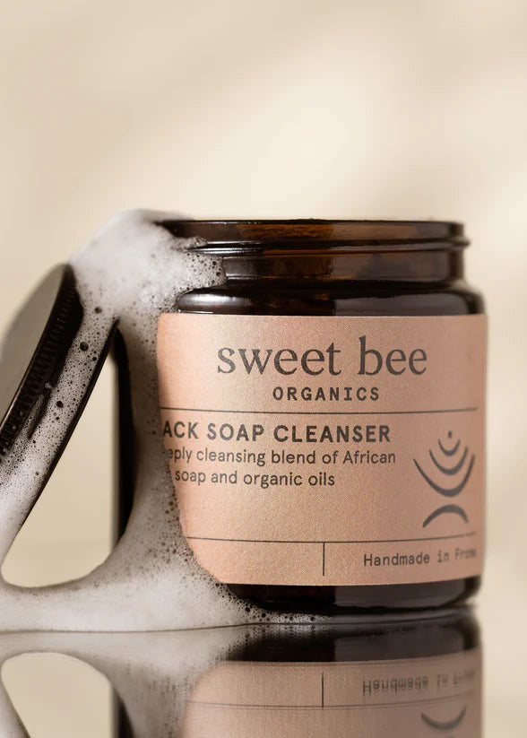 Jar of sweet bee Organics Black Soap Cleanser with a sponge and soap suds on a beige background