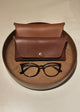 Brown leather case with a button and black glasses on a beige surface