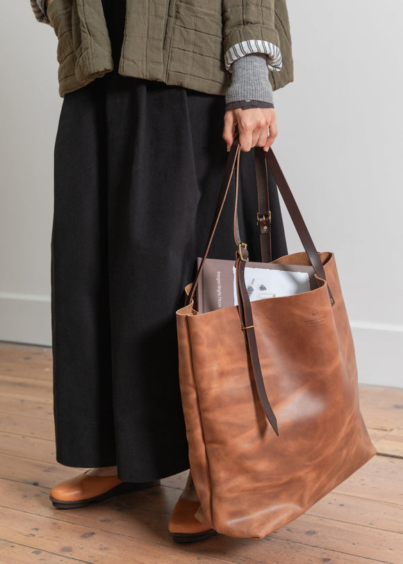 Person holding a brown leather tote bag on a wooden floor.