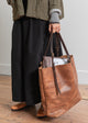 Person holding a brown leather tote bag on a wooden floor.