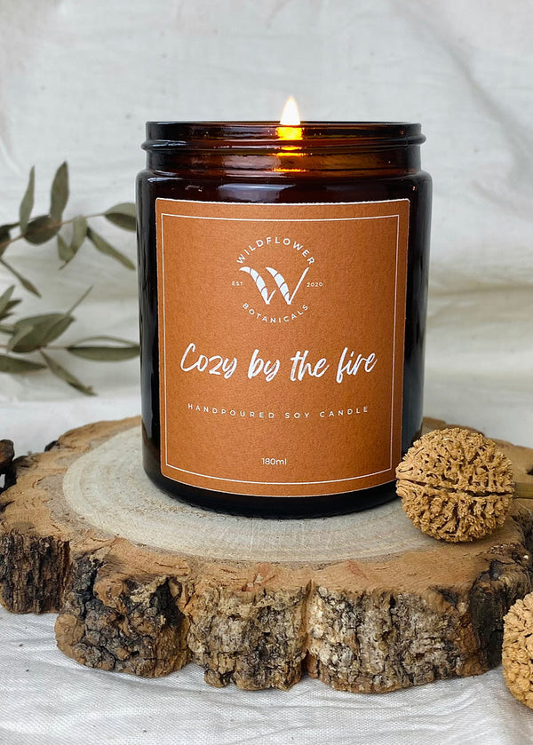 A soy wax candle in a brown jar with a label reading 'Cozy by the fire', surrounded by decorative wildflower botanicals.