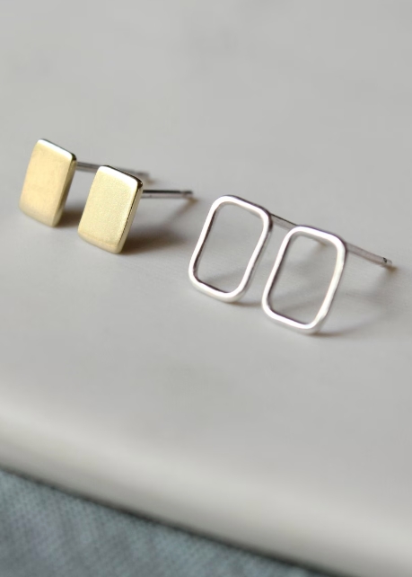 A set of minimalist stud earrings, with one pair in silver and one pair in brass, displayed on a grey surface.