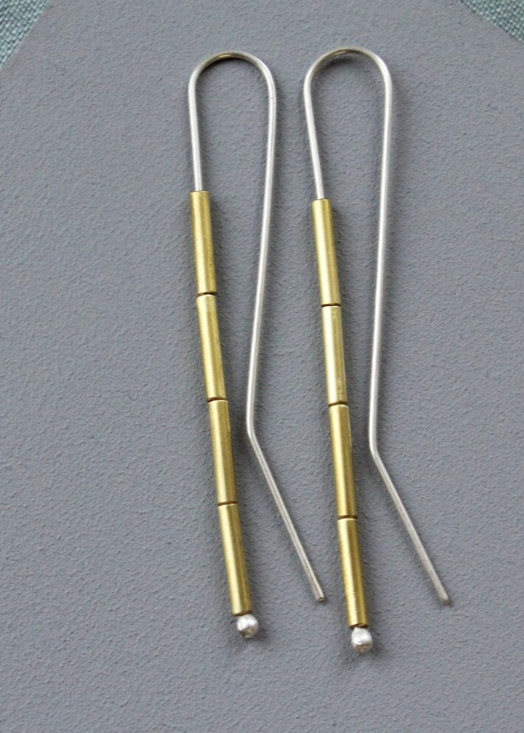 A pair of long brass tube earrings laid on a grey surface, showcasing the silver and gold-colored tubes and the silver ear wire.