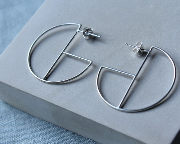 A pair of small geometric silver hoop earrings displayed on a grey surface.