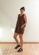 Woman wearing a brown outfit standing on a wooden floor with a white wall background