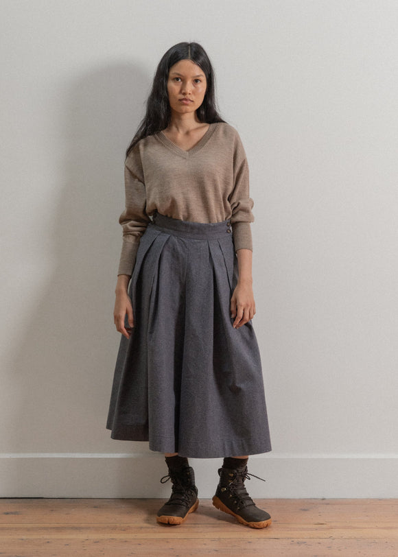 Woman wearing a brown sweater and gray skirt against a plain wall.