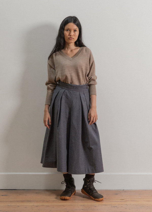 Woman wearing a brown sweater and gray skirt against a plain wall.