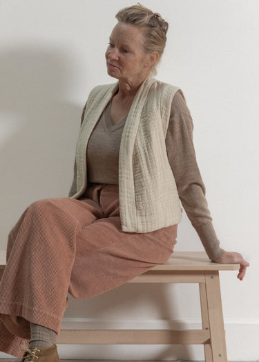Woman sitting on a wooden stool wearing a beige cardigan and brown pants against a white wall.
