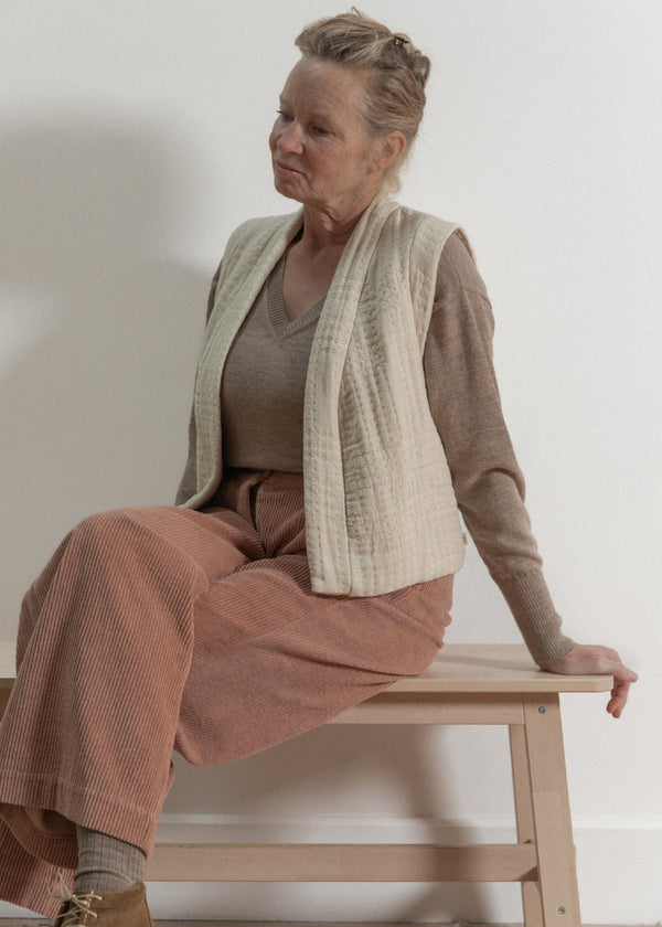 Woman sitting on a wooden stool wearing a beige cardigan and brown pants against a white wall.