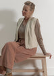 Woman sitting on a wooden stool wearing a beige cardigan and brown pants against a white wall.
