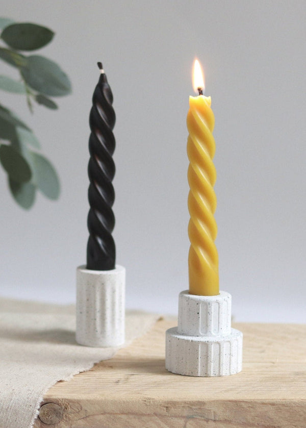 Two twisted candles, one black and one yellow, in white holders on a wooden surface with a light background.