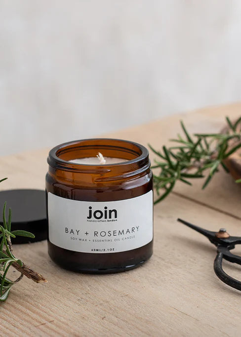 A scented candle with a label reading 'Join', a sprig of rosemary, and a pair of scissors on a wooden surface.