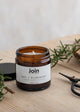 A scented candle with a label reading 'Join', a sprig of rosemary, and a pair of scissors on a wooden surface.