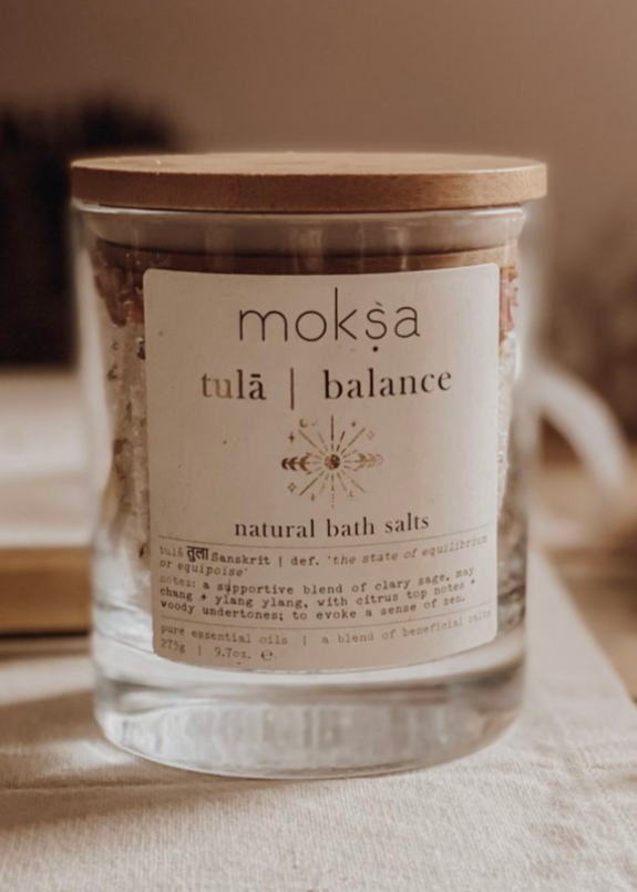 A clear glass jar with a wooden lid labeled 'moksa tula balance' containing natural bath salts, alongside a wooden scoop, on a white towel with a blurred background.
