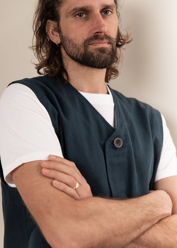 Man wearing a dark vest over a white shirt with arms crossed against a neutral background