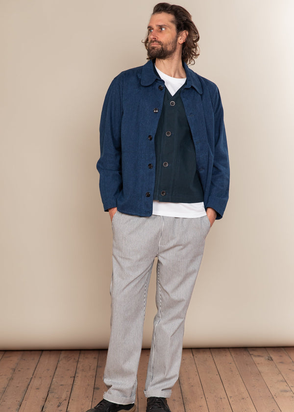 Man wearing a blue jacket, white shirt, and gray pants against a beige wall.